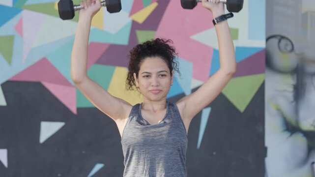 South Asian female athlete performs standing dumbbell shoulder presses during functional fitness training in a modern indoor gym. Mid close shot. Collection: WORKOUTSERIES2026.