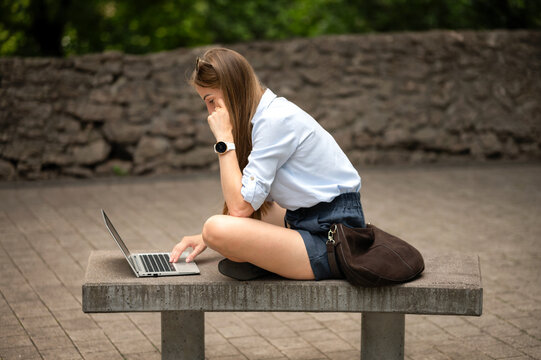 Young female student sitting cross legged on concrete bench and typing on laptop outdoors. Remote education and online learning in city park