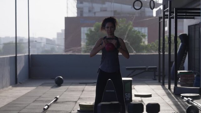 Wide front-angle shot of a fit young woman doing kettlebell goblet sumo squats for leg exercise in an open-air rooftop gym facility under daylight.