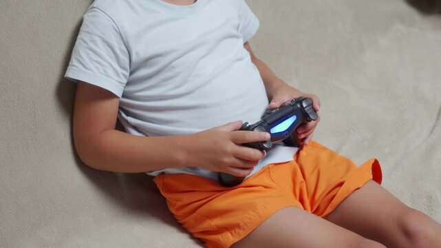 An excited child passionately holds a glowing black gamepad while playing video games. They sit cross-legged and focused on a comfortable, light-colored couch during a fun leisure routine at home.