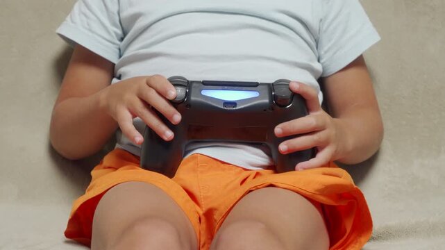 An excited child passionately holds a glowing black gamepad while playing video games. They sit cross-legged and focused on a comfortable, light-colored couch during a fun leisure routine at home.