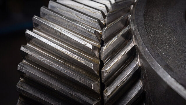 Gears mesh together in a close-up display of heavy industrial machinery featuring metallic textures and sharp geometric teeth under dim ambient lighting.