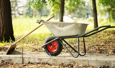 A silver construction wheelbarrow with gloves rests on a newly poured concrete path next to a shovel during park landscaping work. © Aigul Sabirova