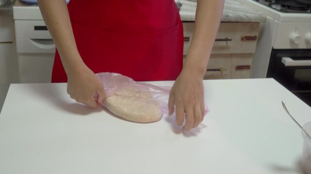 A focused person carefully wraps raw dough in a clear plastic bag to rest it. They work passionately at a clean white table inside a modern kitchen while preparing a delicious homemade meal.