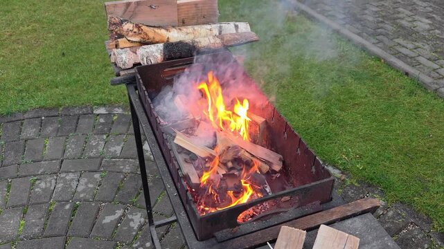 On a tiled area near a grassy lawn there is a metal brazier in which wood chips and firewood burn and smoke comes out. There are birch firewood nearby. Overcast autumn weather