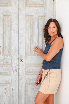A woman stands beside weathered wooden doors with peeling paint, wearing a casual summer outfit in bright sunlight.