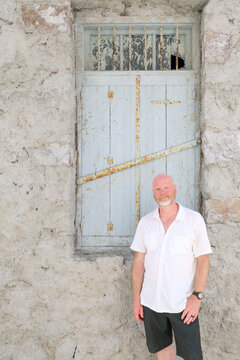 A man stands in front of a weathered stone wall with a rustic shuttered window, bathed in bright sunlight.