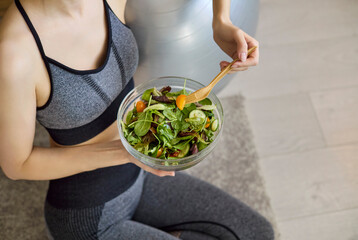 Fototapeta premium Woman eating salad after fitness at home. Athletic woman in sportswear holds a fresh salad bowl, promoting healthy diet and nutrition for wellness content. Concept of clean eating and fitness.