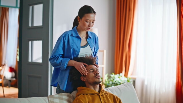 Asian woman giving head massage to boyfriend on sofa. Multiracial couple home.
