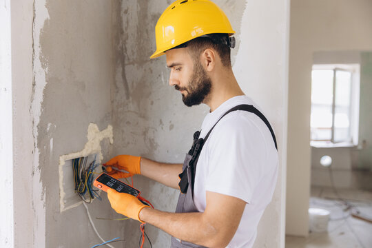 Electrician using a multimeter to check electrical connections and wires in a wall, wearing safety gloves and hard hat