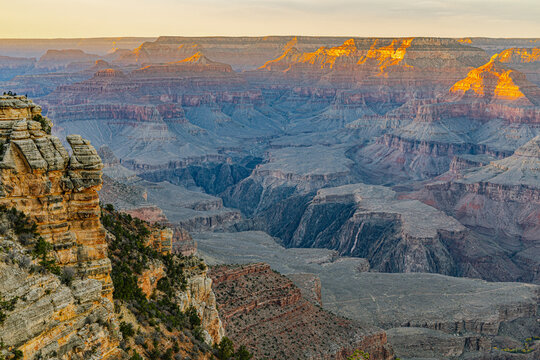 Golden hour light rakes across the Grand Canyon's layered walls and buttes, viewed from a rocky South Rim overlook.
