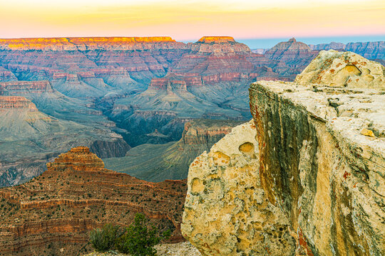A weathered rock ledge frames a sweeping sunrise view over the Grand Canyon's layered cliffs and buttes, Arizona.
