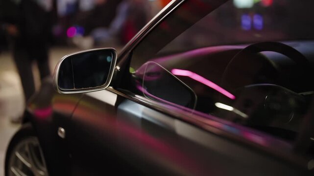 Modern black car parked on a city street at night with vibrant neon lights reflecting off its glossy surface, showing a close-up view of the side mirror and window in slow motion