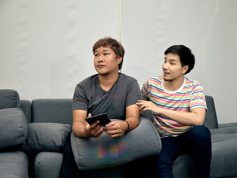 Two men sitting on a sofa watching television, one holding a remote while the other offers support, showing friendship, concern, conversation, comfort, empathy, companionship, and home relaxation