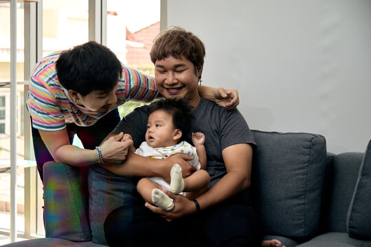 Two smiling men caring for a baby on a sofa at home, sharing love, affection, bonding, support, happiness, comfort, tenderness, and joyful caregiving together