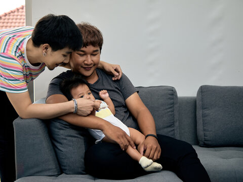 Two smiling men caring for a baby on a sofa at home, sharing affection, family bonding, parenthood, tenderness, happiness, comfort, support, love, and togetherness