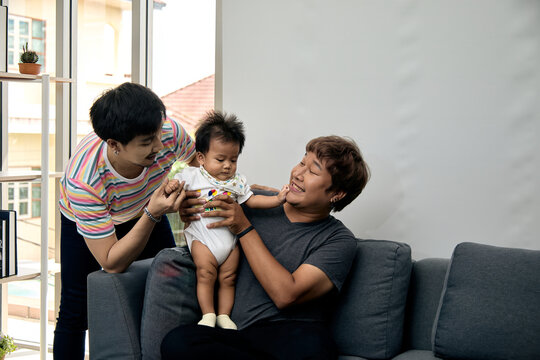 Two smiling men supporting a standing baby on a sofa at home, sharing family care, affection, bonding, happiness, tenderness, parenthood, love, comfort, and joyful togetherness