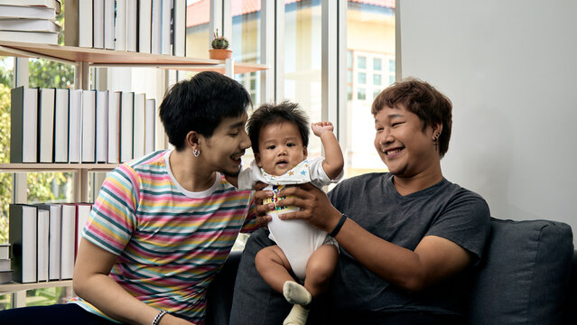 Two smiling men holding a baby on a sofa at home, sharing happiness, care, affection, bonding, support, comfort, love, and joyful togetherness in a bright living room