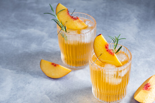 Two glasses hold refreshing drinks with ice and peach slices with a sprig of rosemary on a gray background. Herbs are added for flavor. The scene is bright and set on a light surface.