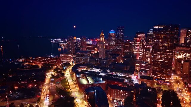 Brightly-lit streets of Boston, Massachusetts, USA at night. Footage over the luminous city along the waterfront of the river. The Moon in the dark sky over the scenery.