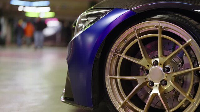 Purple customized luxury sports car with matte finish, carbon fiber details and golden alloy wheels parked indoors at a motor show, showing the front fender, tire, and headlight