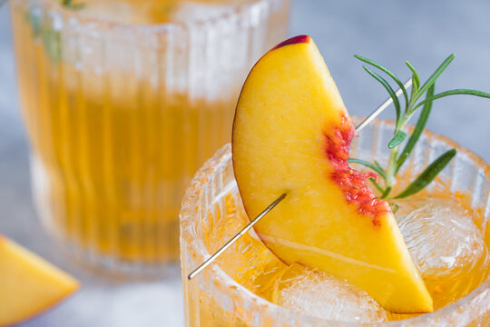 A close view shows a cocktail with ice cubes in a glass. A peach slice and a sprig of herbs decorate the drink. This scene takes place in a bar during daytime.