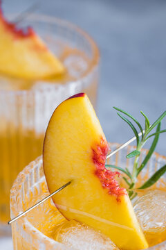 A close view shows a cocktail with ice cubes in a glass. A peach slice and a sprig of herbs decorate the drink. This scene takes place in a bar during daytime.