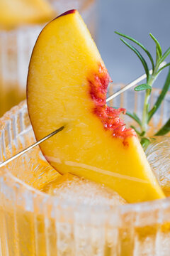 A close view shows a cocktail with ice cubes in a glass. A peach slice and a sprig of herbs decorate the drink. This scene takes place in a bar during daytime.