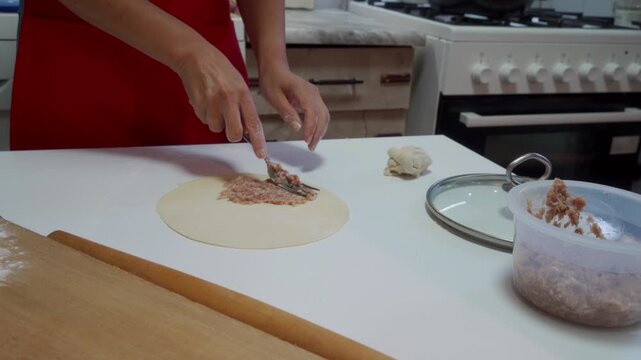 A woman places meat filling onto a circular piece of dough with a fork on a white kitchen table. She feels focused and passionate while carefully preparing food during her home cooking routine.
