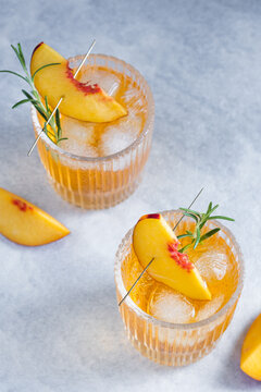 Two glasses hold refreshing drinks with ice and peach slices with a sprig of rosemary on a gray background. Herbs are added for flavor. The scene is bright and set on a light surface.