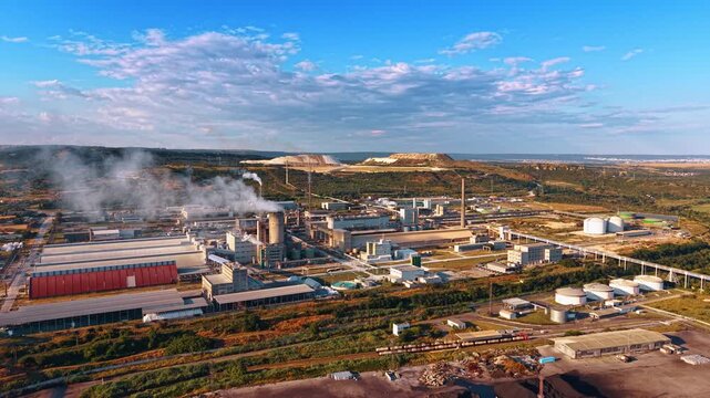 Industrial plant with smoking chimney. An aerial wide shot shows the manufacturing facility surrounded by green hills under a bright cloudy sky.