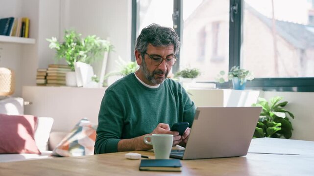 Video of mature man working with laptop while using smartphone at home