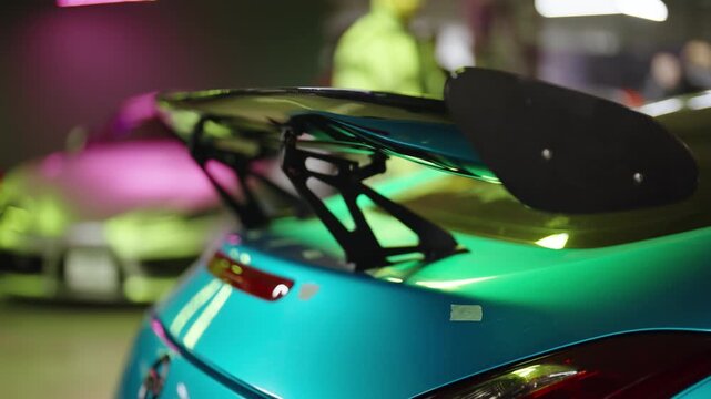 Close-up view of a custom turquoise sports car with a large black spoiler, gleaming under vibrant neon lights at a tuning show, showcasing its sleek, modern, and aerodynamic design