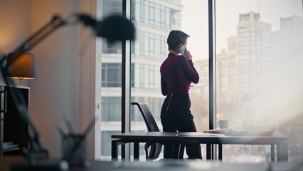 Fototapeta premium Businesswoman talking phone by window office. Faceless lady gazing city skyline.