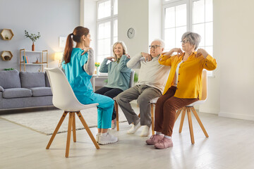 Fototapeta premium Group of smiling elderly people sitting on chairs, doing exercises with young female caregiver in nursing home. Joyful seniors engaging in physical therapy for mobility, wellness and active lifestyle.