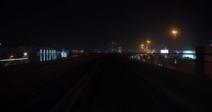Fast POV timelapse from the front window of a driverless metro train speeding along elevated tracks at night as stations and city lights rush past