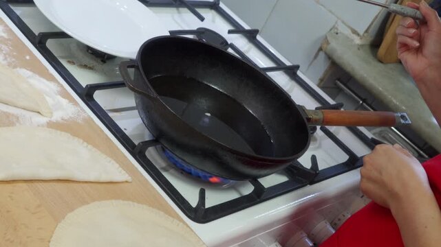 A woman waits as oil heats in a large black pan over a blue gas flame. She feels excited, eager, and hungry while watching the sizzling stove in a cozy kitchen.
