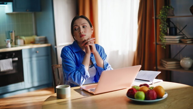 Thoughtful freelancer working laptop home kitchen. Asian girl remote job