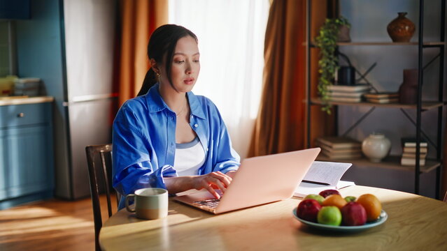 Freelancer typing pink laptop dining table. Asian woman cozy morning routine home