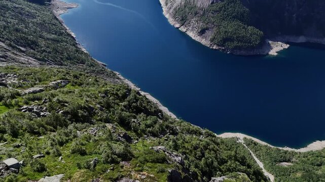 Drone perspective of deep blue fjord bordered by steep forested mountains and rocky shoreline in summer, near Trolltunga in western Norway.