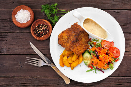 German Jagerschnitzel with a mushroom cream sauce, potato wedges and salad. Top view table scene on a dark wood background.