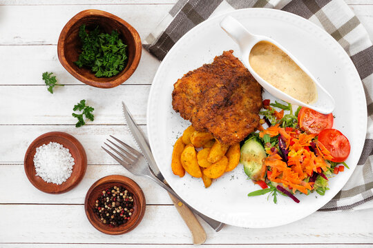 Traditional German Jagerschnitzel with a mushroom cream sauce, potato wedges and salad. Top down view table scene on a white wood background.
