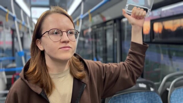A young woman stands in a bus, holding a handrail with determination. The bus's interior shows empty seats and sunlight reflecting through the windows.
