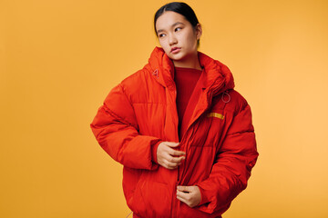 Fototapeta premium Young woman in bright red puffer jacket against solid yellow background, posing with thoughtful expression and casual style for fashion and winter clothing concept.
