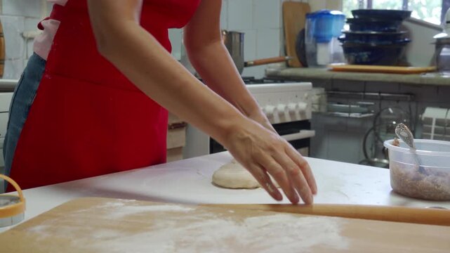 Close-up of woman in red dress rolling dough on heavily floured wooden countertop while preparing pastry in busy home kitchen; visible hands pressing rolling pin over pale dough, scattered flour