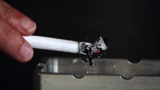 Slow-motion video of an isolated hand against a black background holding a lit cigarette with glowing embers and dropping the ash into an ashtray. Concept of a smoker smoking