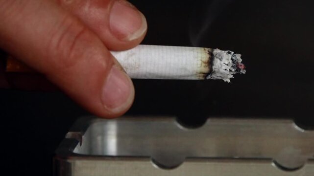 Slow-motion video of an isolated hand against a black background holding a lit cigarette with glowing embers and dropping the ash into an ashtray. Concept of a smoker smoking