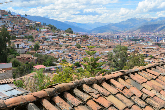 Old terracotta tiled roofs and panoramic view of Cusco, Peru, on Andean slopes, with blue sky and clouds.