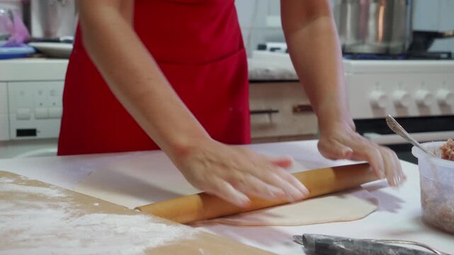 Close-up of adult hands pressing wooden rolling pin over flat dough on flour-covered countertop, with baking tools and stove softly blurred behind, warm natural light creating cozy home cooking mood