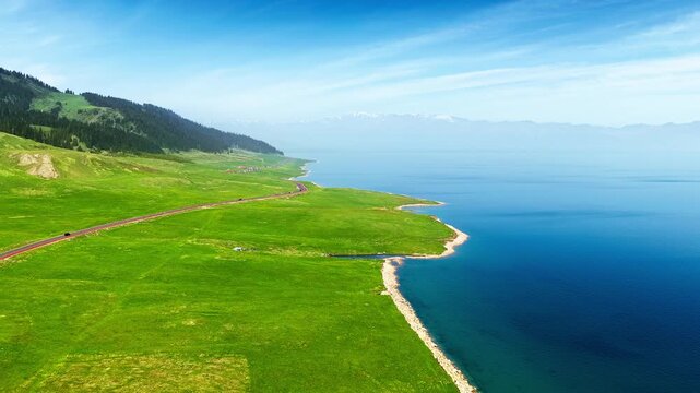 Aerial view of vibrant green grasslands bordering a deep blue lake with distant mountains under a bright blue sky in Xinjiang, China.